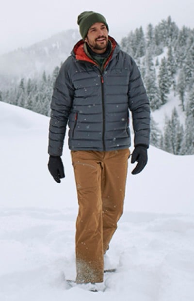 A man in winter clothing walks in snow with trees and a foggy backdrop.