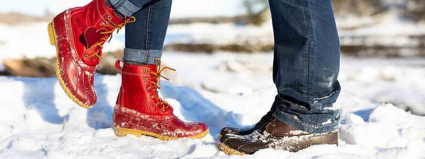 Two sets of Bean Boot clad feet facing each other in the snow, one red Bean Boot raised as if they are kissing.