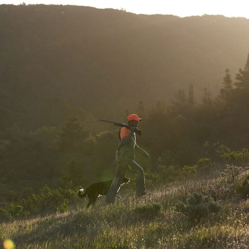 A woman hunter kneeling the field next to a dog, both looking intently into the distance.