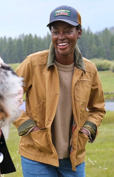 A woman in a jacket and jeans stands outdoors with a background of greenery and water. The scene feels serene and natural.