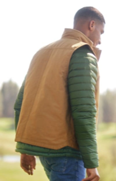 A gentleman is seen from behind wearing a tan vest over a green jacket, with a blurred natural background.