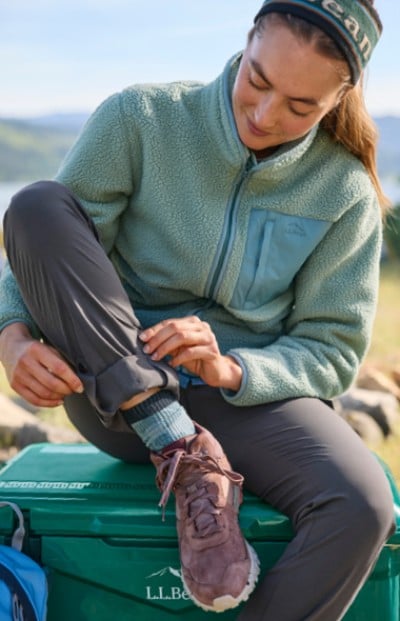 A woman sits on a green cooler outdoors while adjusting her pant leg.