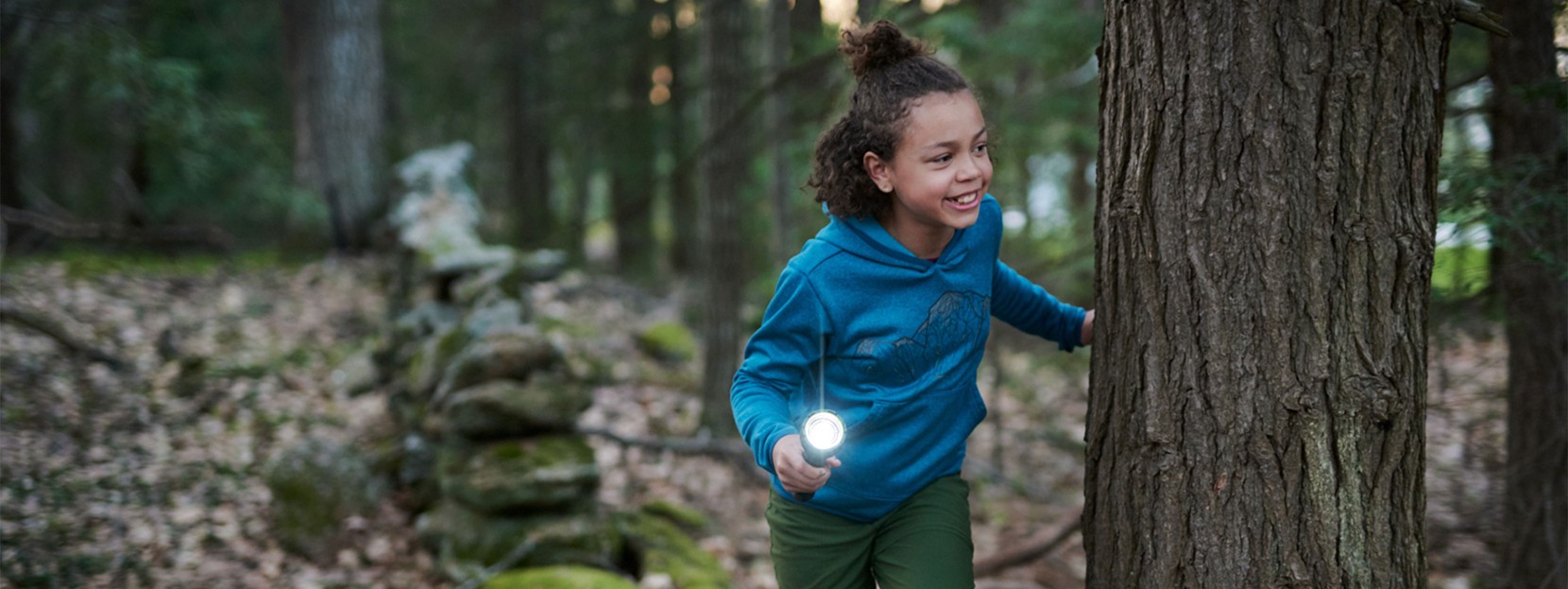 A young girl peeks around a tree with a flashlight playing flashlight tag.