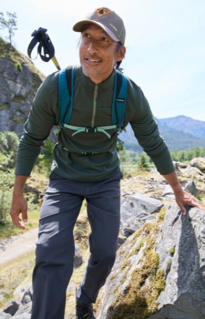 A man climbs rocky terrain with green vegetation, wearing a backpack and holding a hiking pole, with mountains in the background.