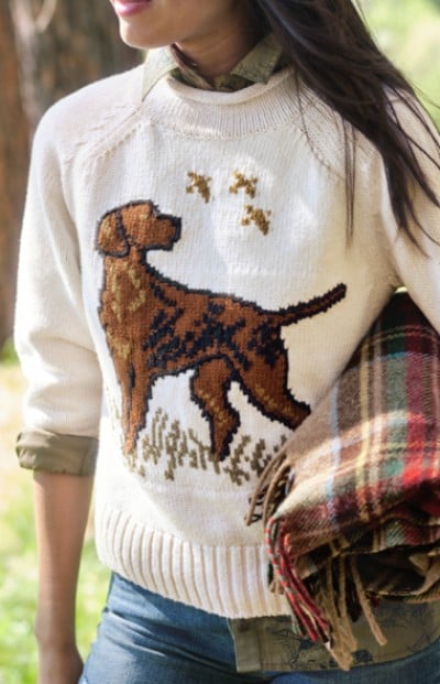 A woman is wearing a sweater with a dog design, standing in a forested area.