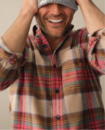 A smiling man waist up, hands on head, wearing a plaid, flannel shirt.