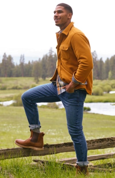 A man is standing on a log in a grassy field with a forest in the background. The scene captures a moment of balance and nature.