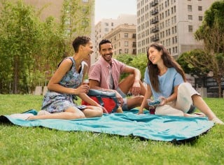 3 friends sitting on a blanket on the grass having a picnic in the city.