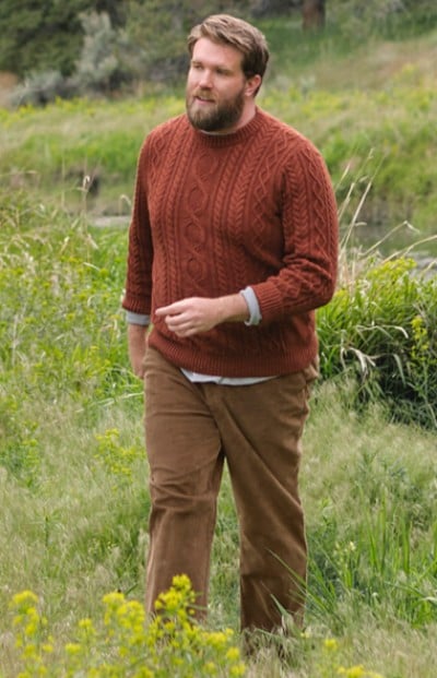 An man in a maroon sweater stands outdoors in a field surrounded by greenery.