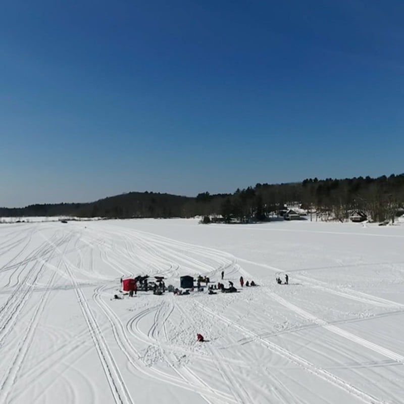 An overhead view from a distance of many people and a few shelters on a snow-covered lake on a beautiful day.