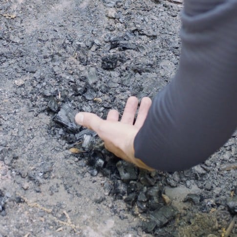 Close-up of Nate's hand being held close to the coals of an extinguished fire.