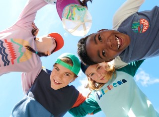 4 smiling kids in a huddle, shot from below looking up at faces and blue sky.