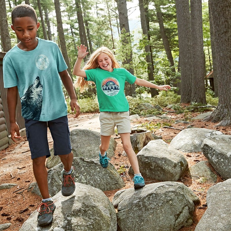 2 happy kids stepping from boulder to boulder on a wooded path.