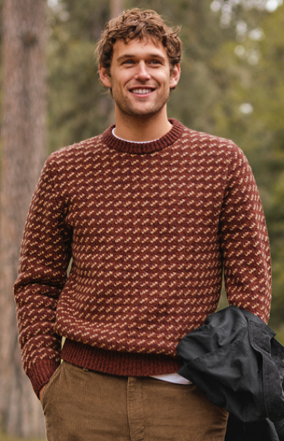 A man stands outdoors, wearing a patterned sweater and holding a jacket. A wooded area surrounds him.