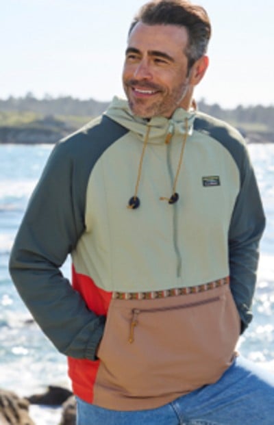 A man stands in front of a wavy body of water, wearing a multi-colored hooded windbreaker.