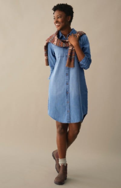 A woman in a denim dress and brown boots stands against a neutral background. The scene is simple and stylish. 